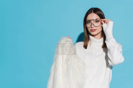 stylish young woman adjusting trendy eyeglasses and holding white faux fur jacket on blue