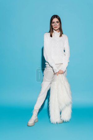 Téléchargez les photos : Toute la longueur de la jeune femme élégante dans des lunettes à la mode et col roulé tenant veste blanche en fausse fourrure sur bleu - en image libre de droit