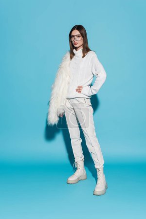 full length of stylish woman in eyeglasses and turtleneck standing with hand on hip while holding faux fur jacket on blue