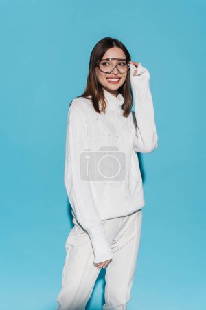 fröhliche junge Frau in trendiger Brille und komplett weißem Outfit posiert auf blauem Grund