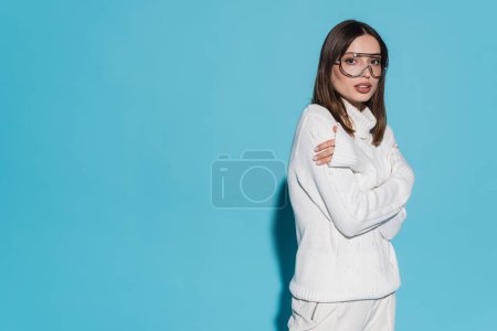 hübsche junge Frau in trendiger Brille und komplett weißem Outfit posiert mit verschränkten Armen auf blauem Grund