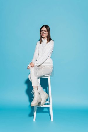 full length of young and stylish woman in total white outfit sitting on high chair on blue
