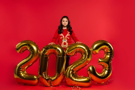 volle Länge des glücklichen Mädchens im Pullover, das neben Luftballons mit 2023 Zahlen auf Rot steht