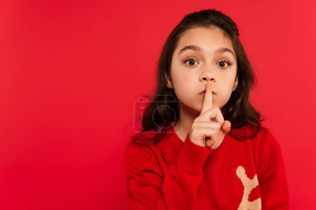 Téléchargez les photos : Enfant en pull d'hiver montrant un signe de silence et regardant la caméra isolée sur rouge - en image libre de droit