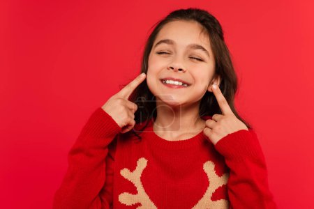 cheerful child in winter sweater pointing with fingers at face isolated on red