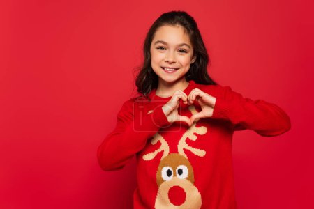 cheerful child in winter sweater showing heart sign with hands isolated on red