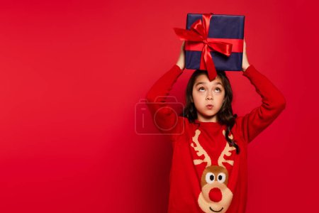 Überraschtes Kind im Winterpullover hält Weihnachtsgeschenk über dem Kopf auf Rot