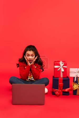 full length of shocked kid in winter sweater and jeans sitting near laptop and Christmas presents on red