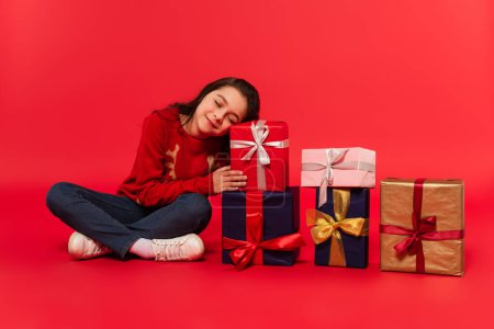 Téléchargez les photos : Longueur complète de l'enfant heureux en pull d'hiver et jeans assis près de cadeaux de Noël sur rouge - en image libre de droit