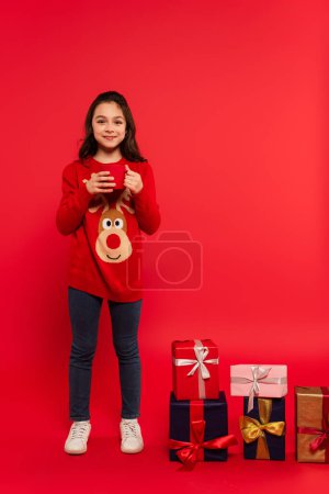 Téléchargez les photos : Pleine longueur de l'enfant heureux en pull tricoté tenant tasse de cacao près de cadeaux de Noël sur rouge - en image libre de droit