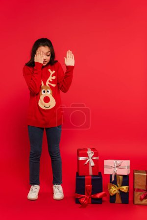 full length of surprised child in knitted sweater looking at Christmas presents on red