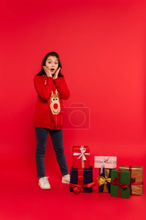 full length of shocked child in knitted sweater near Christmas presents on red