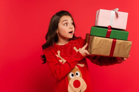 amazed girl in winter sweater holding bunch of Christmas presents and smiling isolated on red