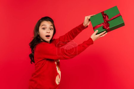 shocked girl in winter sweater holding wrapped Christmas present isolated on red