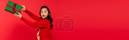 shocked girl in winter sweater holding wrapped Christmas present isolated on red, banner