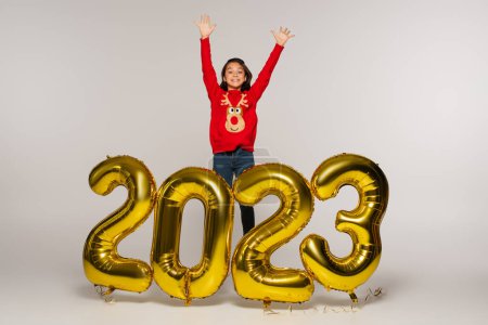Ein fröhliches Kind in rotem Pullover steht mit erhobenen Händen in der Nähe von Luftballons mit 2023 Ziffern auf grau