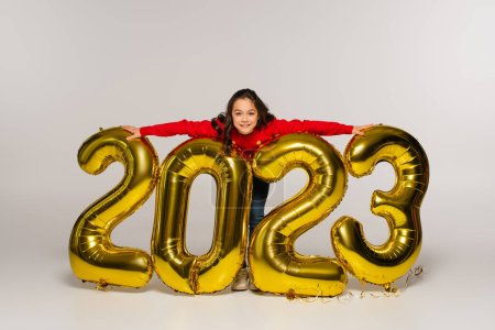full length of cheerful kid in red sweater hugging balloons with 2023 numbers on grey