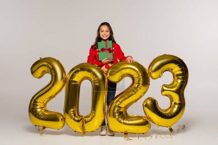 full length of happy kid in  sweater holding christmas present near balloons with 2023 numbers on grey