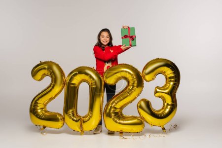 full length of cheerful kid in red sweater holding present near balloons with 2023 numbers on grey