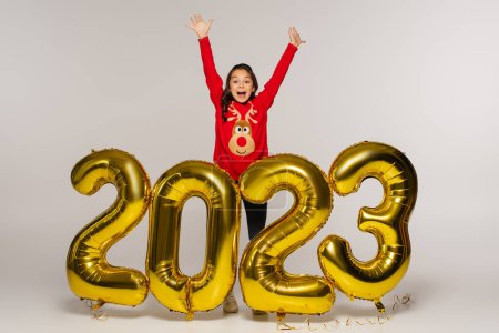 full length of amazed kid in red sweater standing with raised hands near balloons with 2023 numbers on grey