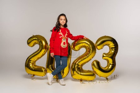 Ein fröhliches Kind in rotem Pullover steht neben Luftballons mit 2023 Ziffern auf grau