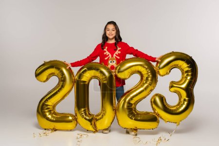 in voller Länge lächelndes Mädchen in rotem Pullover, das neben Luftballons mit 2023 Zahlen auf grau steht