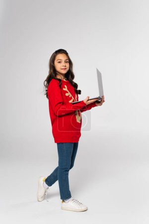 full length of smiling child in red Christmas sweater holding laptop on grey