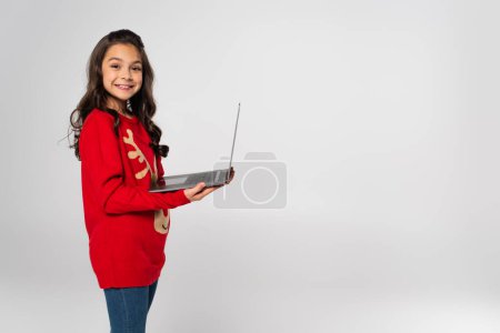 happy girl in red Christmas sweater holding laptop isolated on grey