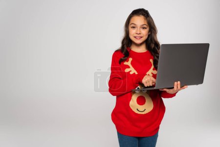 Lächelndes Kind im roten Weihnachtspullover mit Laptop auf grauem Untergrund