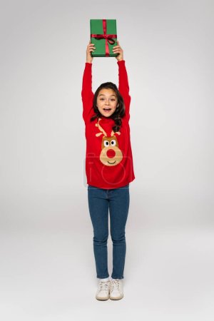 full length of excited girl in red sweater holding Christmas present above head on grey