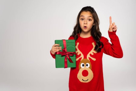 surprised girl in red sweater holding wrapped Christmas present and pointing with finger isolated on grey