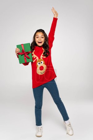 full length of amazed girl in red sweater holding Christmas present on grey