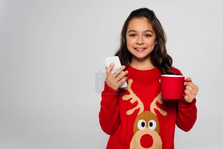 Positive child in New Year sweater holding cup and smartphone isolated on grey 