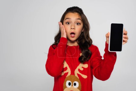 Shocked girl in Christmas sweater holding smartphone with blank screen isolated on grey 
