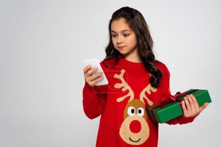 Preteen girl in Christmas sweater holding gift and using smartphone isolated on grey 