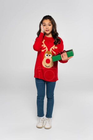 Full length of girl in sweater talking on smartphone and holding Christmas present on grey  