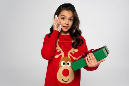 Preteen girl in sweater talking on smartphone and holding Christmas gift isolated on grey 