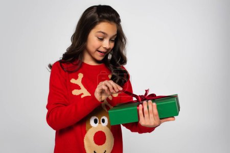 Aufgeregtes Kind im Pullover hält Weihnachtsgeschenk mit Schleife auf Grau 
