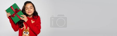 Smiling girl in sweater holding Christmas gift isolated on grey, banner 