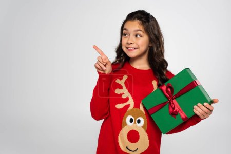 Smiling girl in festive sweater holding gift and pointing with finger isolated on grey 