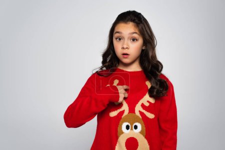 Shocked girl in festive red sweater pointing with finger isolated on grey 
