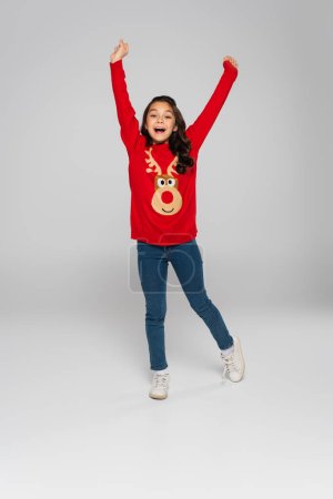 Foto de Longitud completa de niño alegre en suéter de punto rojo mirando a la cámara sobre fondo gris - Imagen libre de derechos