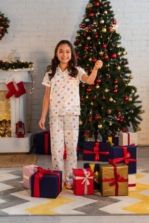 Lächelndes Kind im Pyjama hält Wunderkerze in der Nähe von Geschenken und Weihnachtsbaum zu Hause 