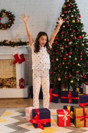 Aufgeregtes Mädchen im gepunkteten Pyjama schaut zu Hause auf Geschenkschachteln neben dem Weihnachtsbaum 