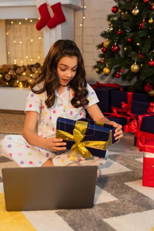 Excited kid in dotted pajama opening gift box near laptop and Christmas tree at home 
