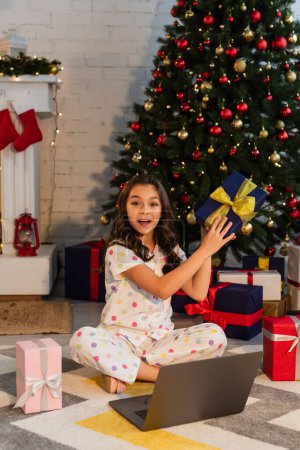 Excited girl in pajama holding present and looking at camera near laptop and Christmas tree at home 