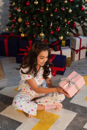 Cheerful kid in dotted pajama holding gift box near blurred Christmas tree at home 