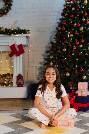 Hübsches Kind im Pyjama blickt in die Kamera, während es neben dem Weihnachtsbaum sitzt und abends zu Hause Geschenke verteilt 