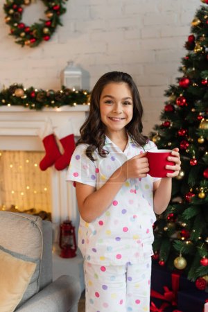 Positives Kind im Pyjama hält abends Tasse neben Weihnachtsbaum zu Hause 
