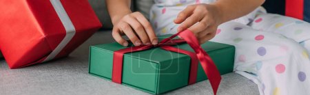 Téléchargez les photos : Vue recadrée de l'enfant en pyjama déliant arc sur le cadeau de Noël sur le canapé, bannière - en image libre de droit